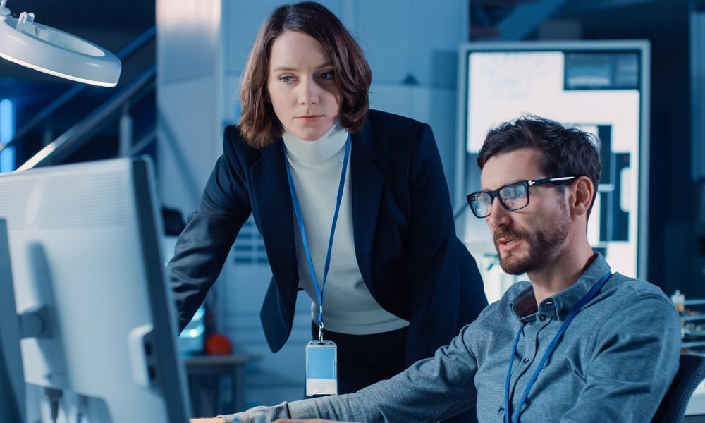 Futuristic Machine Engine Development Engineer Working On Computer At His Desk, Talks With Female Project Manager. Team Of Professionals Working In The Modern Industrial Design Institution